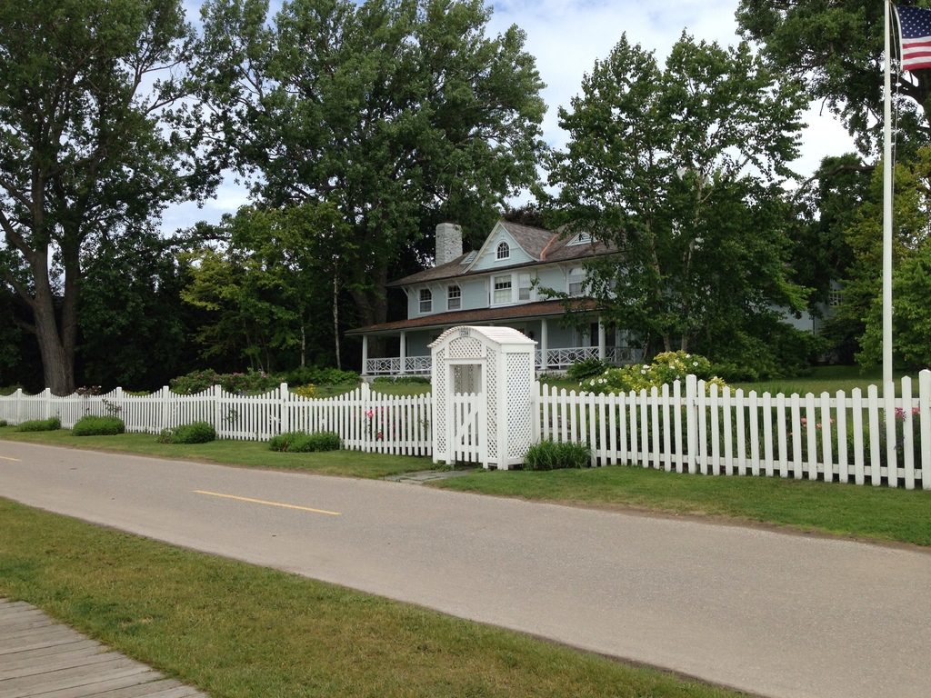 A white farmhouse with a wraparound porch behind a white picket fence and tall trees