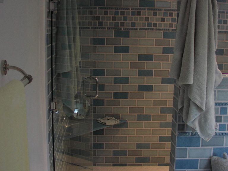 A walk in shower lined with blue and grey brick tiles behind a clear glass panel